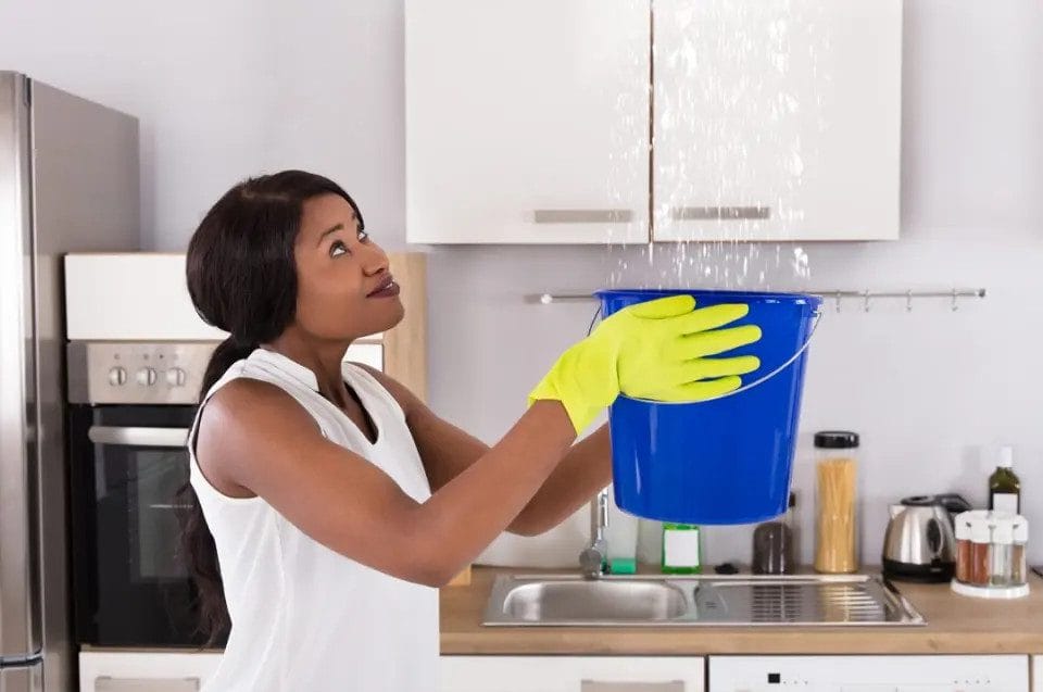 A woman stands in her kitchen, holding a bucket of water and catching water dripping from the ceiling.